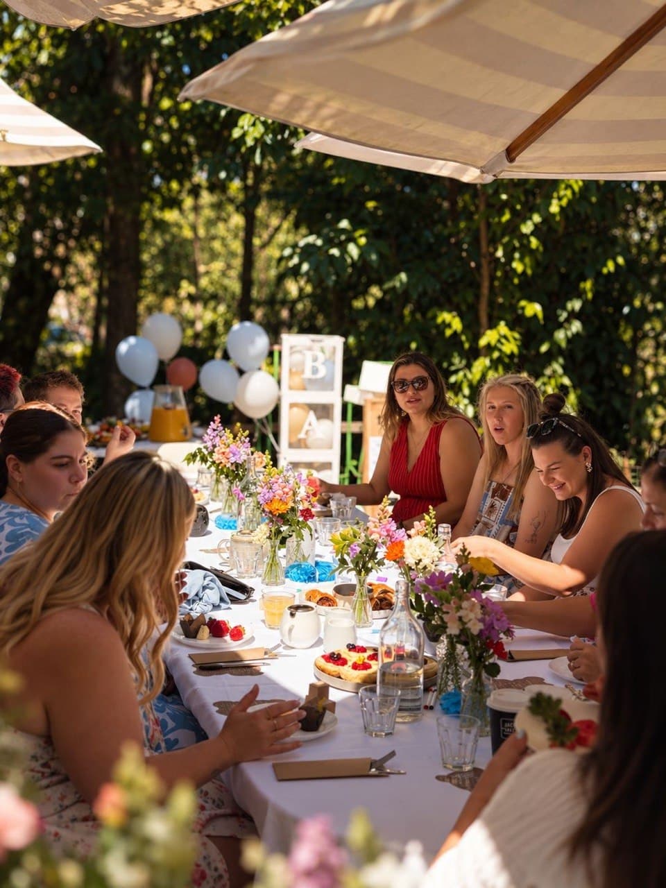 Guests at a long table under striped umbrellas for a baby shower in the Fairhill gardens, Sunshine Coast, with flowers, pastries and balloons