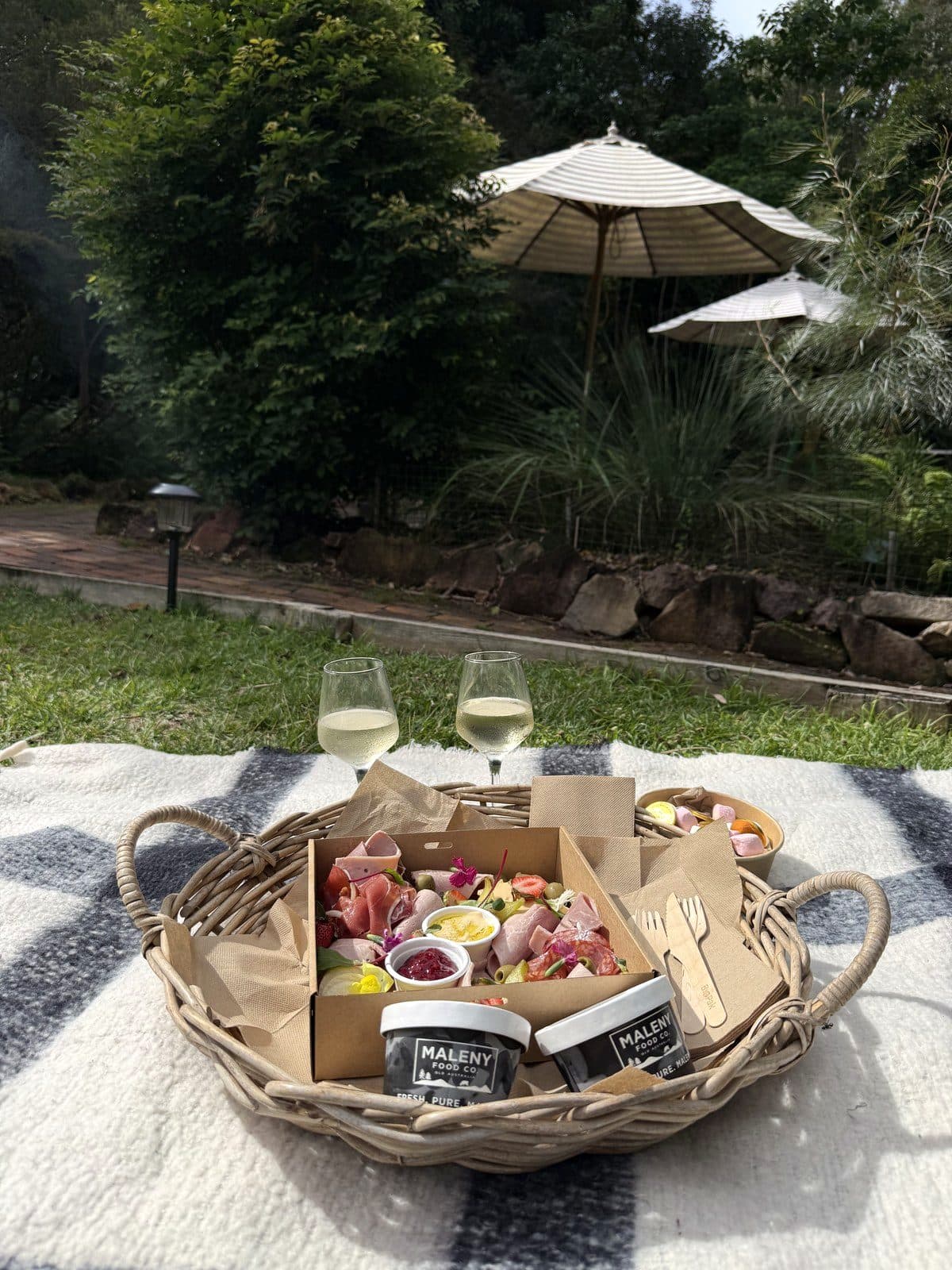 A picnic basket with bubbles on a rug in the Fairhill gardens, sun umbrellas beyond
