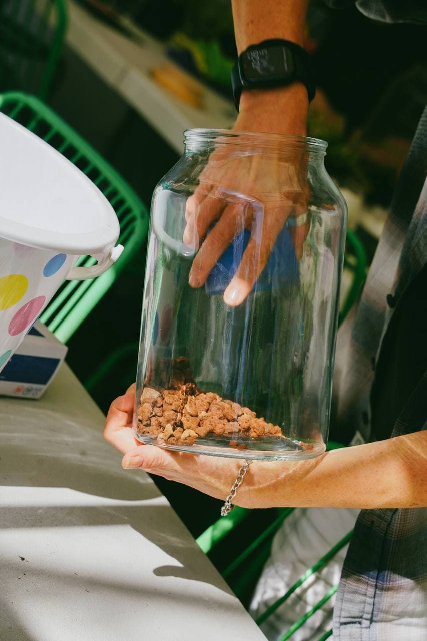 Hands layering pebbles into the base of a glass terrarium jar
