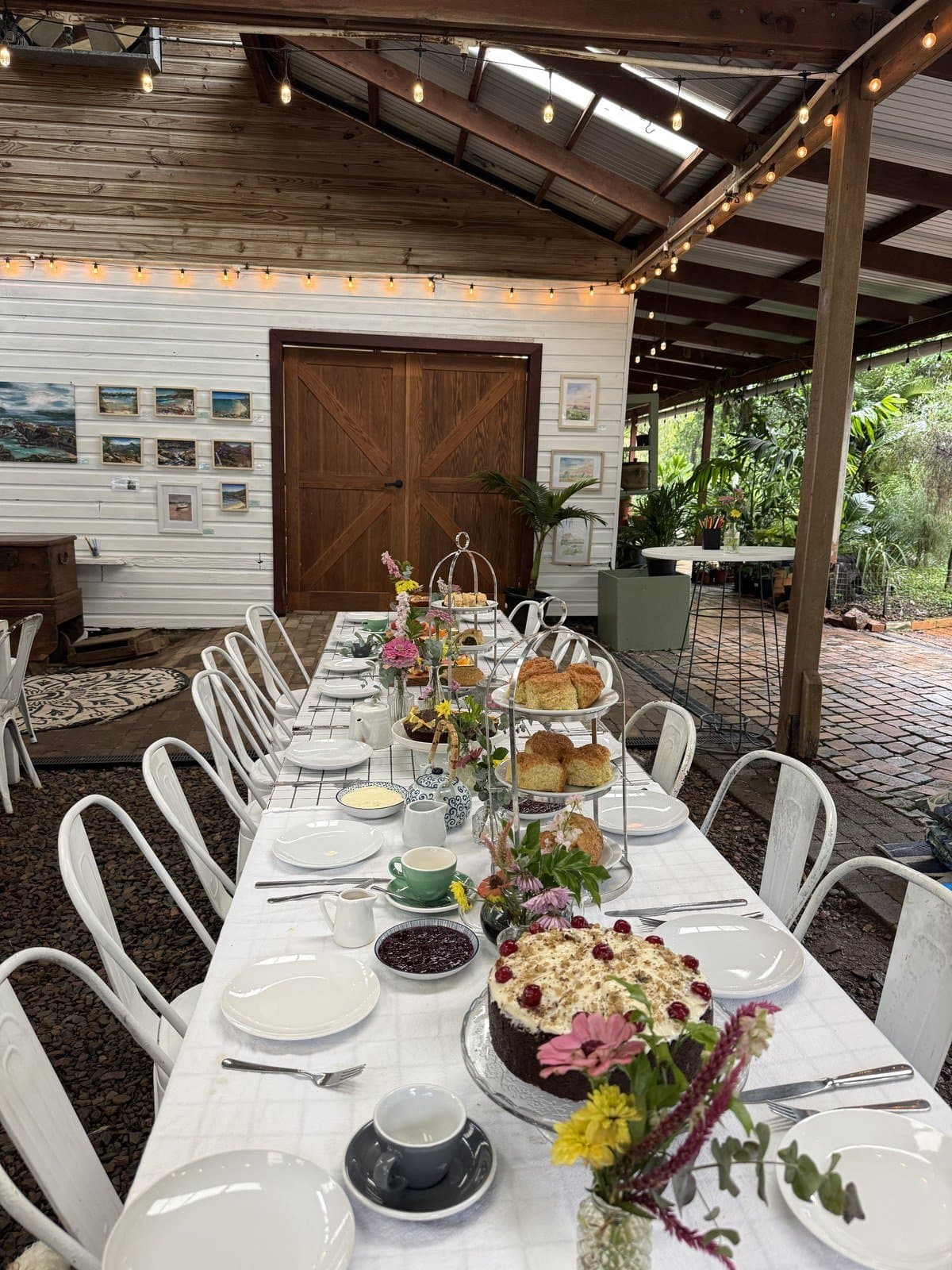 A long morning tea table set under fairy lights in the Fairhill Pavilion, with scone stands and flowers down its length