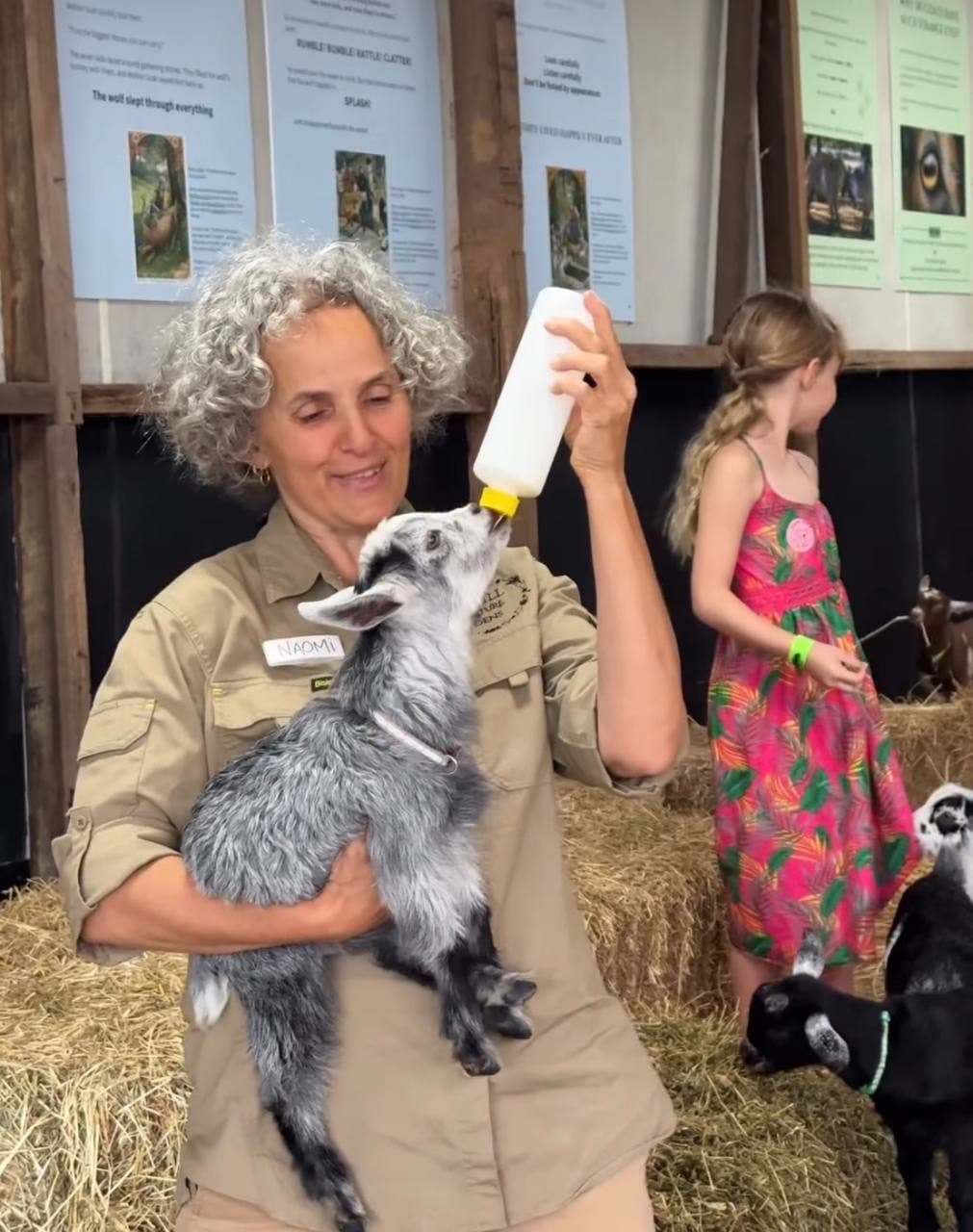 A Fairhill Ranger bottle-feeding a goat kid beside a young guest in the Playbarn